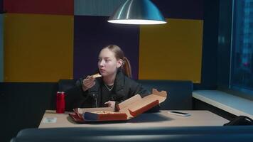 Blonde haired young woman casually dining in colorful restaurant, enjoying takeaway pizza while sipping soda and browsing smartphone during evening break video