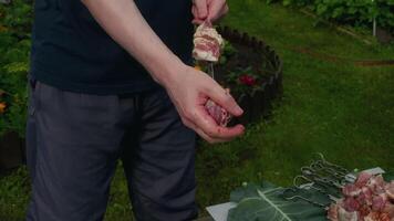 Cook preparing delicious pork and bacon skewers in the garden, expertly sliding seasoned meat onto metal skewers with hands and knife, creating a mouthwatering outdoor meal video