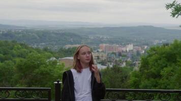 Thoughtful teenage girl with hand in hair standing by railing at viewpoint. Female tourist is exploring city during vacation. Beautiful scenery of buildings amidst green landscape in background. video