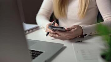 fechar acima do mulher mãos segurando telefone e ativamente usando isto. ela pergaminhos através em formação e escreve baixa importante em formação dentro dela caderno. Móvel telefone é ferramenta para obtendo qualquer Informação. video