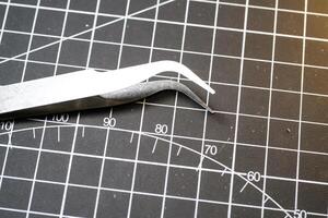 Metal precision tweezer with pointed tip. Tweezer is on a black cutting mat with lines. A tool that functions to clamp and pull out small objects. photo