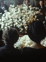 Two women in black hats sitting in front of a large white flower arrangement photo