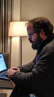 A man with glasses and a beard sitting at a table with a laptop photo