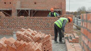 Construction workers building brick wall. Two construction workers in safety gear build a brick wall at a construction site, with tools and stacked bricks visible. video