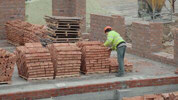 Worker organizing bricks at construction site. A construction worker in safety gear arranges stacks of bricks on wooden pallets at an active building site under development. video