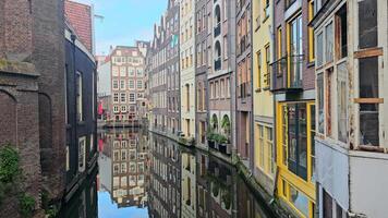 Historic canal in Amsterdam with colorful, narrow buildings reflected in calm water. The scene showcases classic Dutch architecture, with flags and plants adding charm to the urban landscape video