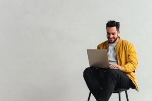 Man in yellow jacket enjoys working on laptop while seated in modern office chair with a light gray background photo