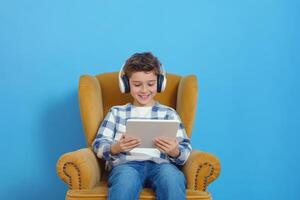 Joyful young boy immersed in reading on a tablet while sitting comfortably in an armchair against a vibrant blue backdrop photo