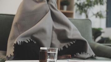 A woman with a blanket on her lap sitting on a couch with a glass of water and a video