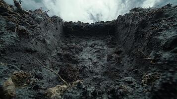 dramatic perspective captures freshly dug burial pit revealing dark soil walls against bright sky. Faint roots and silhouette edge enhance its eerie atmosphere photo