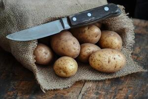Raw potatoes spilling from burlap sack with knife on rustic wooden table photo