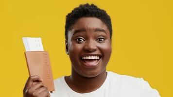 A young African American woman with a plump figure is smiling widely while holding a passport in her hand against a bright yellow background. She shows excitement and joy. video