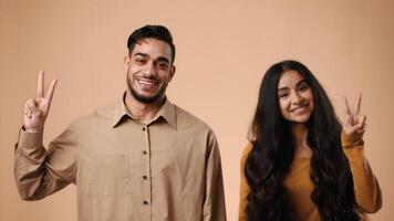 A young Arab couple stands in a studio with a neutral background, smiling and making peace signs with their fingers. They exude joy and warmth in their poses together. video