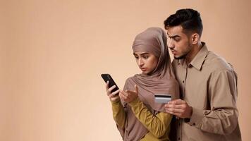 A young Middle Eastern couple stands together in a studio, exploring online shopping on a smartphone. The husband holds a credit card while the wife scrolls through options. video