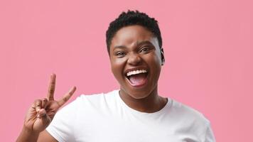 A joyful young African American woman with a chubby figure is smiling widely and giving a peace sign. She is in a studio setting with a vibrant pink background, exuding positivity and confidence. video