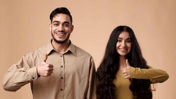 A young married couple from the Middle East stands together against a neutral backdrop, smiling and giving thumbs up, showcasing their joy and connection in a studio setting. video