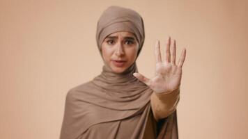 A young Middle Eastern woman wearing a hijab stands against a plain background, raising her hand in a stop gesture to convey a message. She appears focused and serious in her expression. video