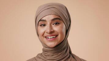 A young Arab woman, wearing a hijab, is smiling warmly in a neutral studio setting. The soft background enhances her cheerful expression and presents a positive atmosphere. video