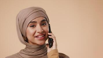 A young Arab woman with a hijab expresses joy while engaged in conversation on her smartphone. She stands in a simple studio environment with a neutral background. video