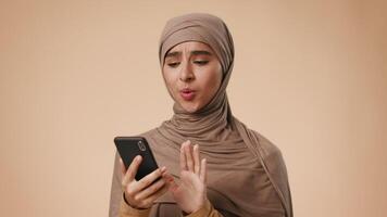 A young Middle Eastern woman, draped in a beige hijab, appears focused while using her smartphone. She is in a studio with a plain background, isolated and absorbed in her device. video