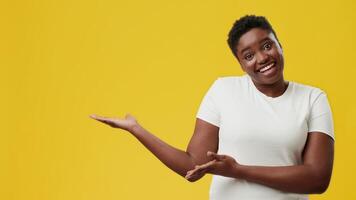 A joyful young African American woman poses with a cheerful expression, gesturing with her hands against a bright yellow background. She exudes confidence and positivity in a studio setting. video