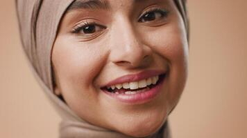 A young Arab woman with a warm smile is posing confidently against a neutral studio background. She is wearing a hijab, radiating positivity and charm in this inviting atmosphere. video
