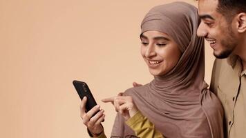 A young Arab couple stands close together in a studio with a warm background. The woman wears a hijab and smiles as she shows something on her phone to her husband, who looks intrigued. video