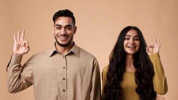 A young Arab couple stands together in a studio setting, smiling at the camera. They are showing a hand gesture that signifies happiness or approval, engaging with each other in a moment of joy. video