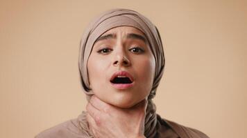 A young Arab woman with a hijab displays a surprised expression while gripping her neck in a studio against a neutral background. This striking image captures an intense moment of emotion. video