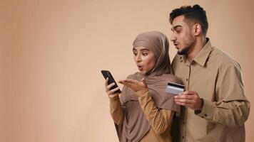 A young Middle Eastern couple is focused on their smartphone while discussing an online purchase. The man holds a credit card, preparing to complete the transaction. video