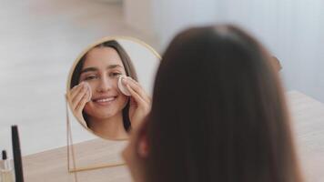 uma milenar árabe mulher é mimos ela mesma com uma cuidados com a pele rotina às lar. ela sorrisos enquanto aplicando produtos para dela face dentro frente do uma circular espelho. video