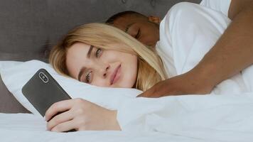 A young multiracial couple cuddles in bed, sharing a tender moment. The woman looks at her phone with a smile while the man embraces her from behind, creating a warm atmosphere. video