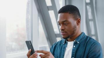 A young African American man in casual attire focuses on his smartphone while waiting at the airport. He appears engaged and attentive as he navigates through his device. video