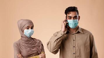 A married Middle Eastern couple stands in a studio against a neutral background. The woman, dressed in modest attire, appears concerned while the man talks on the phone, both wearing face masks. video