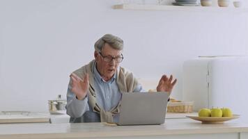 A retired elderly man in his 60s sits at a kitchen table, using a laptop for online shopping. He holds a credit card and appears focused, enjoying his retirement time at home. video