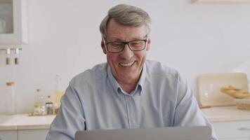 An elderly man in his 60s sits at a kitchen table, focused on his laptop. He wears glasses and casually dressed, surrounded by a tidy home environment. His expression shows concentration. video