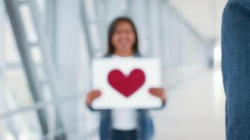 A young African American woman holds a heart-shaped sign at the airport, while a man turns to face her. They share a moment of joy amidst the bustling environment of travelers. video