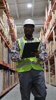 African American warehouse employee in a hard hat and safety vest walking through a storage aisle, inspecting inventory and checking items on a clipboard video