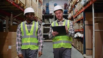 Two warehouse workers in reflective vests and hard hats are discussing tasks. They collaborate efficiently in the organized aisles of a busy warehouse video