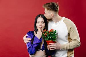 Shocked lady and positive man holding pot with red flowers, looking at camera isolated on blue background. photo