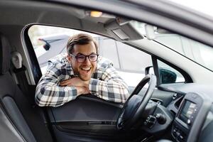 Young man looking through window inside new car, checking it before purchase at modern dealership store. Cheerful guy buying or renting automobile at showroom. photo