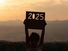 Silhouette of cute little girl holding 2025 sign on sunset background. Happy New Year 2025. photo