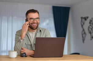 A smiling man talking on his phone and using a laptop while working at home. photo