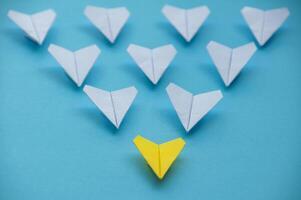 Straight view of yellow paper airplane origami leading other white airplanes on blue background with customizable space for text. Business and Leadership skills concept photo