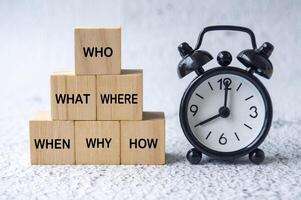Who, What, Where, When, Why and How text on wooden cubes with table clock background. Fact finding concept photo