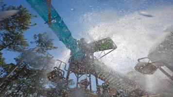 Low-angle view of a colorful water park structure with a bright blue slide, wooden platforms, and buckets spilling water under a clear blue sky surrounded by trees video