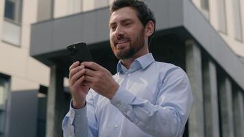 Smiling happy male employer holding mobile phone in city glad satisfied businessman business chat service app company online carefree man guy entrepreneur scrolling using smartphone web net internet video