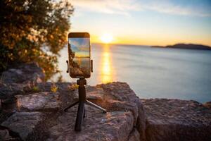 Becici, Montenegro - January 1, 2025. Close-up of an iPhone on a tripod atop a rock, recording a timelapse of the sun setting over the sea. The focus is on the screen capturing the stunning horizon. photo