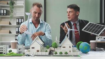 Two men sitting at a table with a model of a house and a wind turbine video