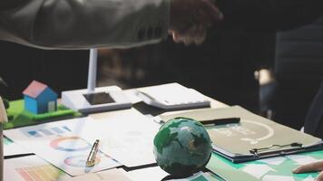 Two people shaking hands over a table with papers and a green globe video
